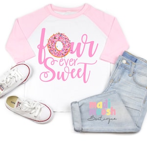 以下が含まれることがあります： ピンクのドーナツのグラフィックと「four ever Sweet」の文字が入った白と薄いピンクのラグランシャツ、 「madi moosh Boutique」の文字が入ったライトブルーのジーンズ、赤と青の縁取りが施された白いローカットのキャンバススニーカー。