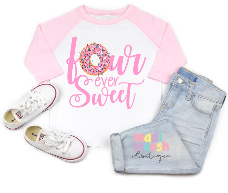 以下が含まれることがあります： ピンクのドーナツのグラフィックと「four ever Sweet」の文字が入った白と薄いピンクのラグランシャツ、 「madi moosh Boutique」の文字が入ったライトブルーのジーンズ、赤と青の縁取りが施された白いローカットのキャンバススニーカー。