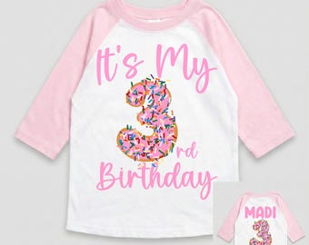 May include: White and pink raglan-style birthday shirt with the text "It's My 3rd Birthday". The number 3 is designed as a pink donut with sprinkles. The back of the shirt has the name "Madi" and a donut.