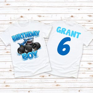 May include: White t-shirts with a shark-themed design. One shirt features a blue shark monster truck with "BIRTHDAY BOY" text. The other shirt has the name "GRANT" and the number "6" in blue.