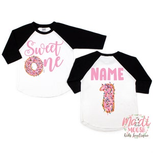 May include: Two white and black baseball shirts with pink text. One shirt says "Sweet One" with a pink donut with sprinkles. The other shirt says "NAME" with a pink donut shaped like the number one with sprinkles.