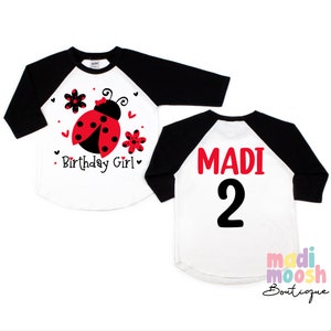 May include: White and black baseball tee shirt with a red ladybug and flowers on the front. The back of the shirt says "MADI 2" in red and black text.