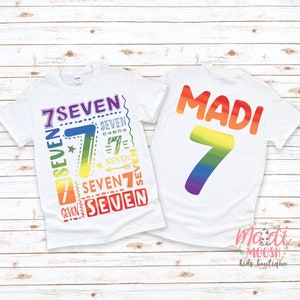 Peut inclure: Deux t-shirts blancs avec des motifs arc-en-ciel. Le t-shirt avant a le chiffre 7 en couleurs arc-en-ciel avec le mot "SEVEN" r&eacute;p&eacute;t&eacute; plusieurs fois. Le t-shirt arri&egrave;re a le nom "MADI" et le chiffre 7 en couleurs arc-en-ciel.