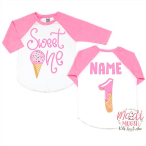 May include: Pink and white baseball tee with the text "Sweet One" and an ice cream cone graphic on the front. The back of the shirt has the text "NAME" and a pink ice cream cone number one graphic.