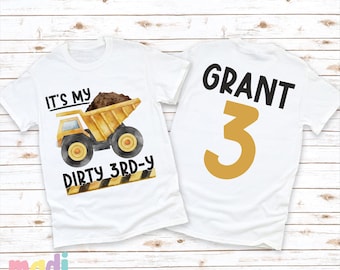 May include: Two white t-shirts with a construction truck theme. The front shirt has a yellow dump truck with the text "It's my dirty 3rd-y". The back shirt has the name "GRANT" and the number "3" in gold.