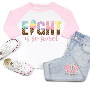 May include: A white and pink baseball tee shirt with the words "Eight is so sweet" in rainbow colors with ice cream cone graphics. The shirt has a pink and blue color block design.