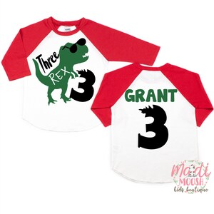 Op de afbeelding: Rood-wit baseballshirt met een groene dinosaurus die een zonnebril draagt en de tekst "Three Rex" op de voorkant. De achterkant van het shirt heeft de naam "GRANT" en het nummer "3" in groen.