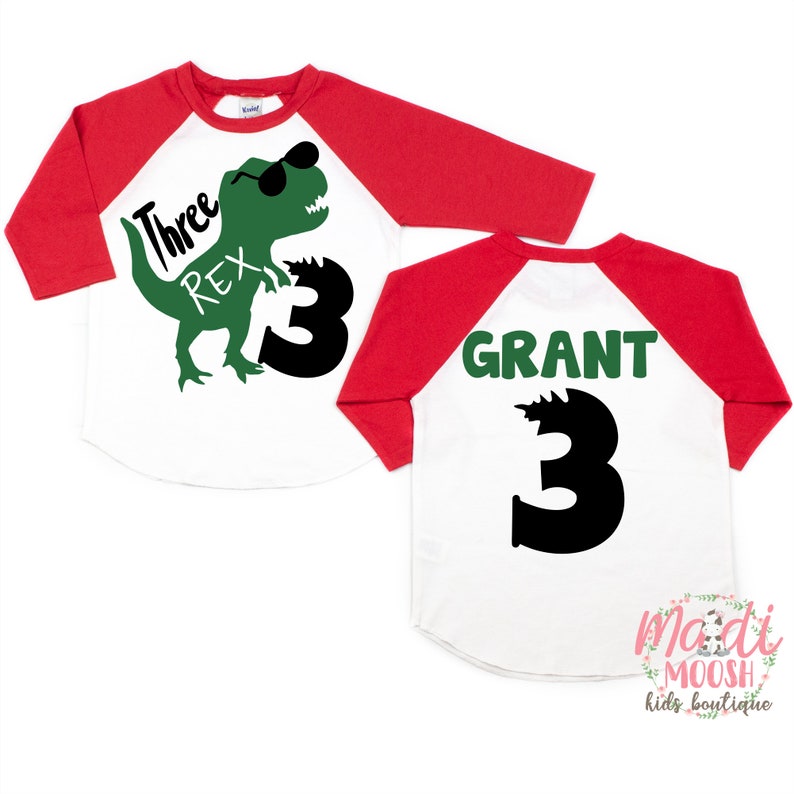 Op de afbeelding: Rood-wit baseballshirt met een groene dinosaurus die een zonnebril draagt en de tekst "Three Rex" op de voorkant. De achterkant van het shirt heeft de naam "GRANT" en het nummer "3" in groen.
