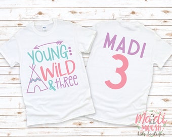 Op de afbeelding: Twee witte T-shirts met een roze, paarse en turquoise ontwerp. De voorkant van een shirt zegt "Young: Wild & Three" met een tipi-afbeelding. De achterkant van het andere shirt zegt "Madi 3".