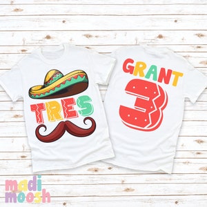 May include: Two white t-shirts with a Mexican fiesta theme. The front shirt has a sombrero, mustache, and the word "TRES" in colorful letters. The back shirt has the name "GRANT" and the number "3" in a colorful, cartoon style.