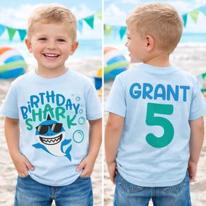 May include: Light blue t-shirt with a shark graphic wearing sunglasses and the words "Birthday Shark". The back of the shirt has the name "Grant" and the number "5" in blue and teal.