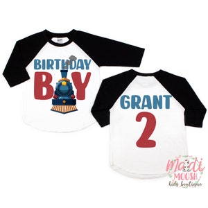 May include: Two white and black baseball shirts with a train design. The front shirt says "Birthday Boy" with a blue and red train. The back shirt says "Grant 2" in blue and red.