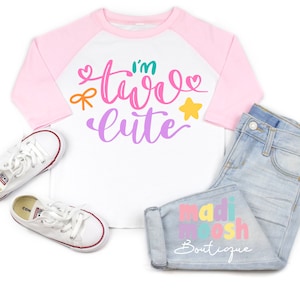 May include: A white and pink toddler t-shirt with the text "I'm two cute" in colorful letters. The shirt has a pink and white baseball style design. The shirt is laying on a white surface with a pair of white sneakers and a pair of blue jeans with the text "madi moosh boutique" in colorful letters.