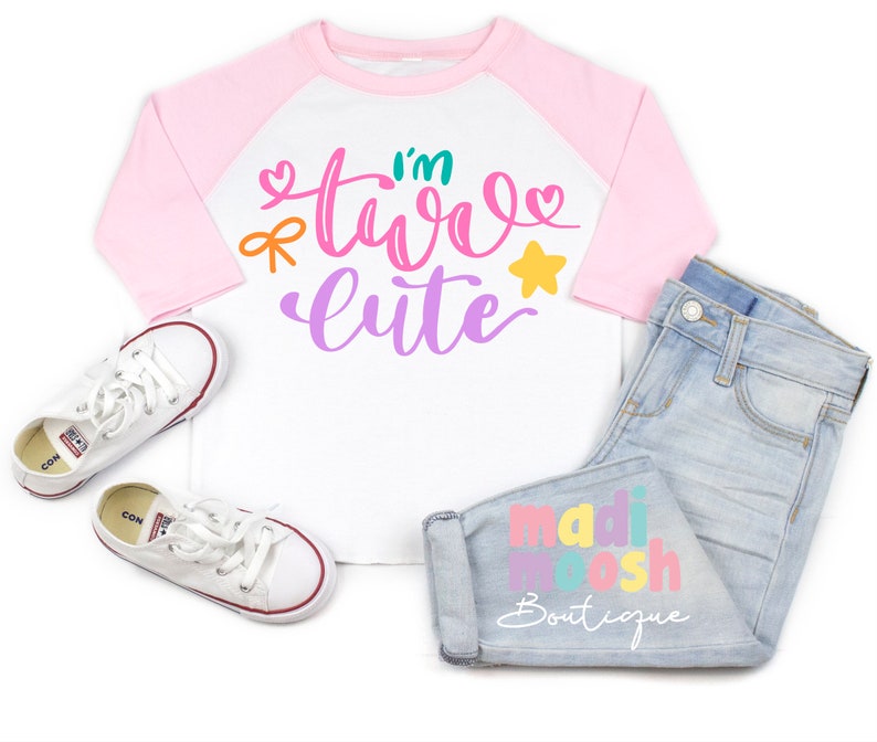 May include: A white and pink toddler t-shirt with the text "I'm two cute" in colorful letters. The shirt has a pink and white baseball style design. The shirt is laying on a white surface with a pair of white sneakers and a pair of blue jeans with the text "madi moosh boutique" in colorful letters.