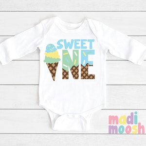 May include: White long-sleeve baby onesie with the text "SWEET ONE" in brown letters. The "ONE" is formed by an ice cream cone with blue, green, and yellow ice cream and sprinkles.