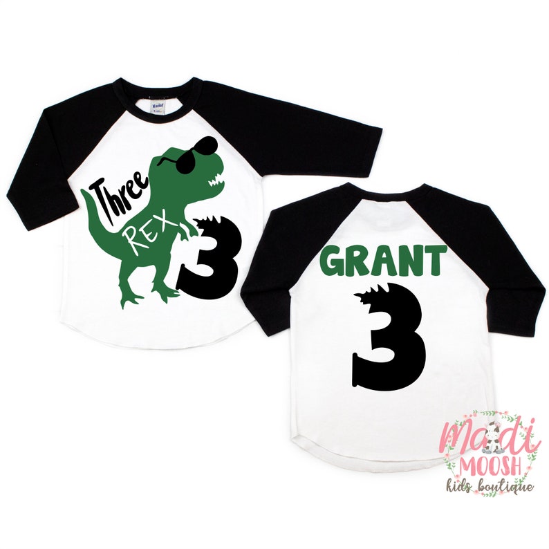 Op de afbeelding: Een wit en zwart baseballshirt met een groene dinosaurus afbeelding en de tekst "Three Rex 3" op de voorkant en de tekst "GRANT 3" op de achterkant.