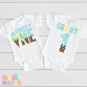 May include: Two white baby onesies with an ice cream cone theme. One onesie says "Sweet One" with a blue and green ice cream cone and sprinkles. The other onesie says "Grant 1" with a blue ice cream cone and sprinkles.
