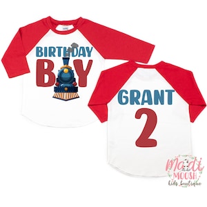 May include: Red and white baseball style shirt with a blue train and the text "Birthday Boy" on the front. The back of the shirt has the text "Grant 2" in blue and red.