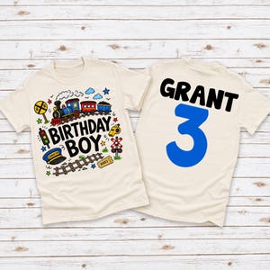 May include: A cream-colored t-shirt featuring a colorful train graphic, railroad tracks, a conductor hat, and the text "BIRTHDAY BOY" on the front, with the name "GRANT" and the large blue number "3" printed on the back of the garment.