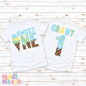 May include: Two white t-shirts with an ice cream cone design. The front shirt says "Sweet One" with the ice cream cone forming the "O" in "One". The back shirt says "Grant 1" with the ice cream cone forming the number "1".