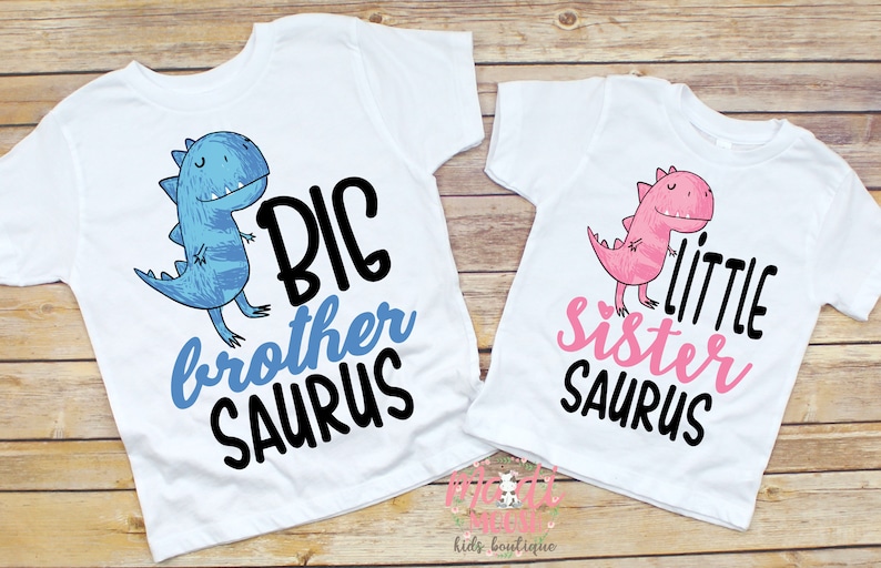 Op de afbeelding: Twee witte T-shirts met dinosaurussen afbeeldingen en tekst. Het blauwe shirt zegt "BIG brother SAURUS" en het roze shirt zegt "little sister SAURUS".