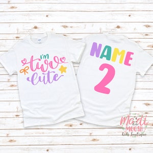 May include: Two white t-shirts with a colorful design. The front shirt says "I'm Two Cute" with a star, bow, and hearts. The back shirt says "NAME 2" in colorful letters. The shirts are on a white wood background.