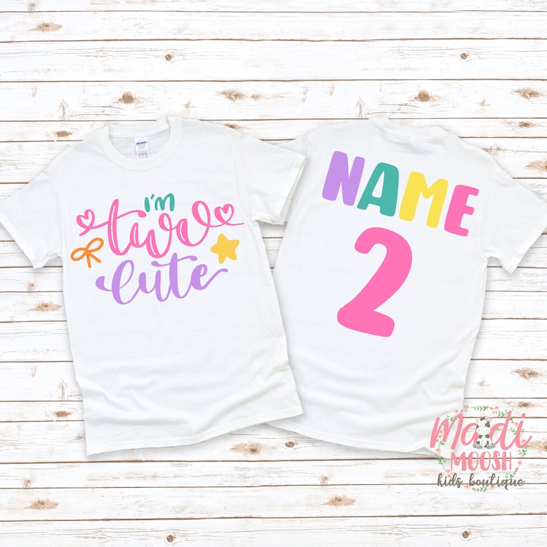 May include: Two white t-shirts with a colorful design. The front shirt says "I'm Two Cute" with a star, bow, and hearts. The back shirt says "NAME 2" in colorful letters. The shirts are on a white wood background.