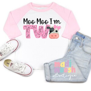 May include: A white and pink raglan t-shirt with the text "Moo Moo I'm Two" in black and pink. A cartoon cow with a pink bow is on the shirt. The shirt is paired with light blue jeans with the text "madi moosh Boutique" in colorful letters.