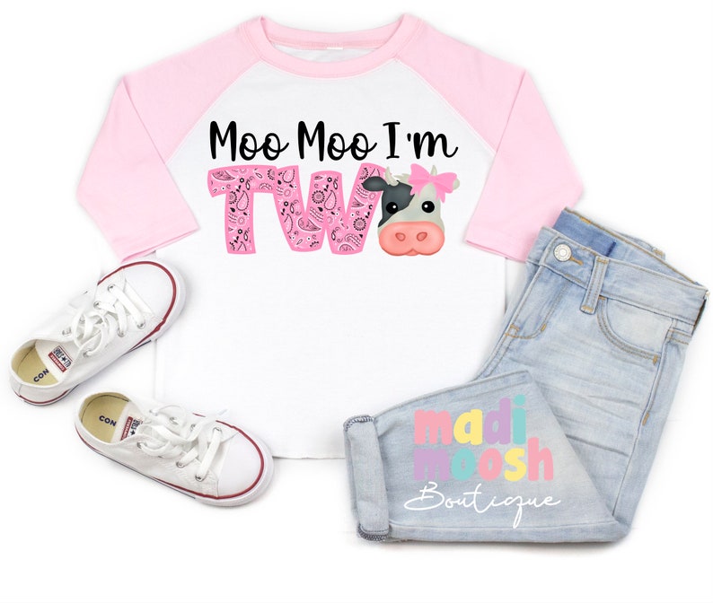 May include: A white and pink raglan t-shirt with the text "Moo Moo I'm Two" in black and pink. A cartoon cow with a pink bow is on the shirt. The shirt is paired with light blue jeans with the text "madi moosh Boutique" in colorful letters.