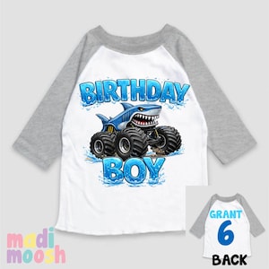 May include: White and gray raglan-sleeve shirt with a blue shark monster truck graphic. The shirt says "BIRTHDAY BOY" in blue text. The back of the shirt has the name "GRANT" and the number "6".