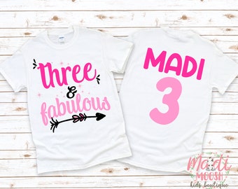 May include: Two white t-shirts with pink and black text. The front shirt says "three & fabulous" with a pink arrow. The back shirt says "MADI 3" with a large pink number 3.