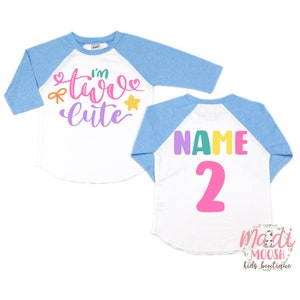 May include: A white and blue baseball style shirt with the text "I'm two cute" in colorful letters on the front and the text "NAME 2" on the back.