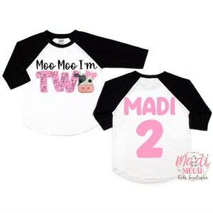 May include: A white and black baseball style shirt with pink lettering that says "Moo Moo I'm Two" with a cow graphic. The back of the shirt says "MADI 2" in pink lettering.