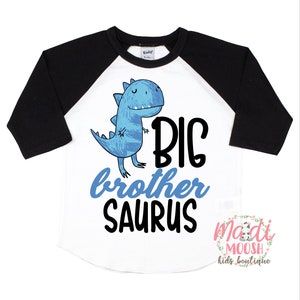 May include: A white and black baseball tee shirt with a blue dinosaur graphic and the text "BIG brother SAURUS".