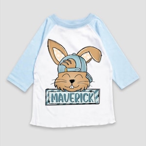 May include: A white and light blue long-sleeved t-shirt with a cartoon bunny wearing a blue baseball cap. The bunny is holding a sign that says "Maverick".