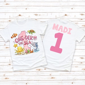 May include: Two white t-shirts. One features the text "ONE-DER THE SEA" with a pink number 1 and sea creature illustrations. The other shirt has the name "MADI" and a pink number 1.