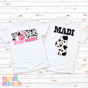 May include: Two white t-shirts with a cow print design. The front of one shirt says "Four Ever Moody" with a pink cow head. The back of the other shirt says "Madi" and has a cow print number "4".