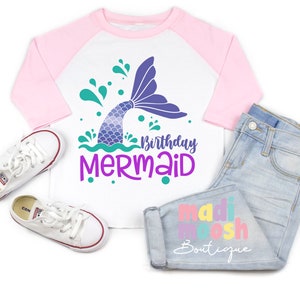 May include: A white and pink long-sleeved shirt with a mermaid tail graphic and the text "Birthday Mermaid". The shirt is on a white background with a pair of white sneakers and blue jeans with the text "madi moosh Boutique" on the leg.