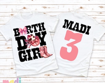 May include: Two white t-shirts with black text. The front shirt says "BIRTHDAY GIRL" with a pink bow, pink cowboy hat, and pink cowboy boots. The back shirt says "MADI 3" with the number 3 in pink.