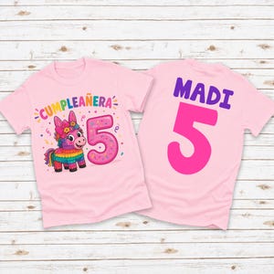 May include: Two pink t-shirts. The front of one shirt has the text "CUMPLEAÑERA" and a colorful piñata with the number "5". The back of the other shirt has the name "MADI" and the number "5".
