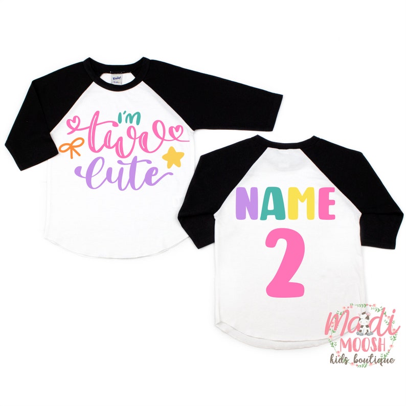 May include: Two-piece white and black baseball tee shirt set for a child's second birthday. The front of the white shirt says "I'm Two Cute" in colorful letters with a heart, bow, and star. The back of the white shirt says "NAME 2" in colorful letters.