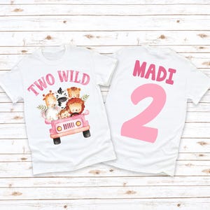 May include: White t-shirts with a pink safari theme. One shirt features the text "TWO WILD" with a watercolor illustration of safari animals in a pink jeep. The other shirt has the name "MADI" and the number "2" in pink.