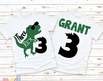 May include: Two white t-shirts with green dinosaur graphics. One shirt says "Three Rex" with a dinosaur wearing sunglasses. The other shirt says "GRANT" with the number "3" in black.