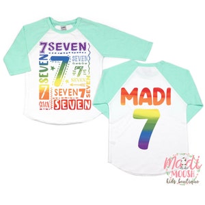 Peut inclure: Un t-shirt de baseball blanc et turquoise avec un motif arc-en-ciel. Le devant du t-shirt porte le chiffre 7 et le mot "seven" r&eacute;p&eacute;t&eacute; plusieurs fois. Le dos du t-shirt porte le nom "Madi" et le chiffre 7 en couleurs arc-en-ciel.