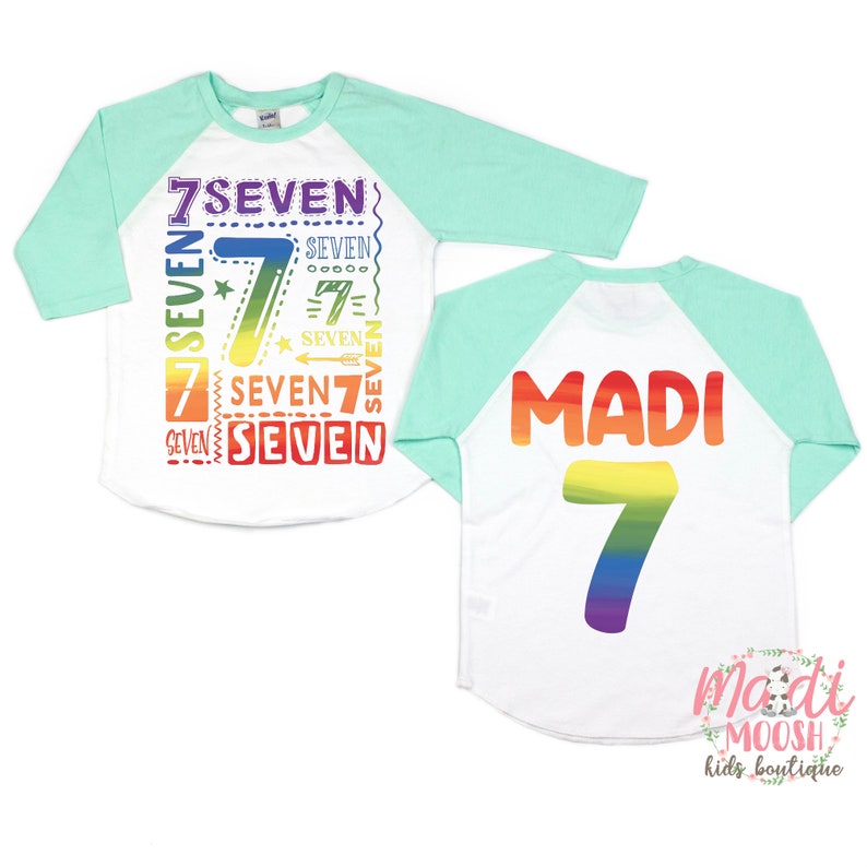 Peut inclure: Un t-shirt de baseball blanc et turquoise avec un motif arc-en-ciel. Le devant du t-shirt porte le chiffre 7 et le mot "seven" r&eacute;p&eacute;t&eacute; plusieurs fois. Le dos du t-shirt porte le nom "Madi" et le chiffre 7 en couleurs arc-en-ciel.