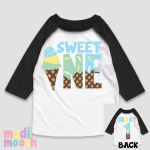 May include: A white and black baseball style shirt with a blue, green, and yellow ice cream cone graphic and the text "SWEET ONE". The back of the shirt has the text "GRANT" and the number "1" in a similar ice cream cone design.