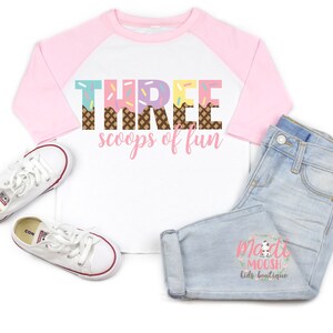 May include: A white and pink baseball tee shirt with the text "THREE scoops of fun" in colorful ice cream cone letters. The shirt has a pink and white color block design.
