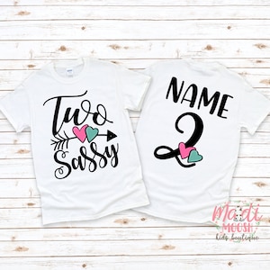 May include: Two white t-shirts with black text and heart graphics. One shirt says "Two Sassy" with an arrow and pink and teal hearts. The other says "Name" and the number "2" with pink and teal hearts.
