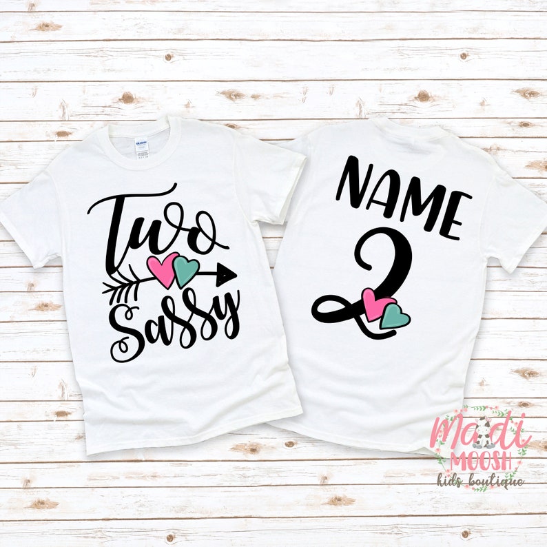 May include: Two white t-shirts with black text and heart graphics. One shirt says "Two Sassy" with an arrow and pink and teal hearts. The other says "Name" and the number "2" with pink and teal hearts.