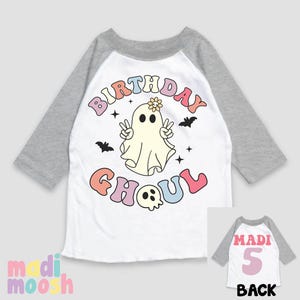 May include: A white and grey raglan t-shirt with a colourful "Birthday Ghoul" graphic featuring a ghost with a flower on its head. The back of the shirt has a pink number "5" and the word "Madi" in pink.